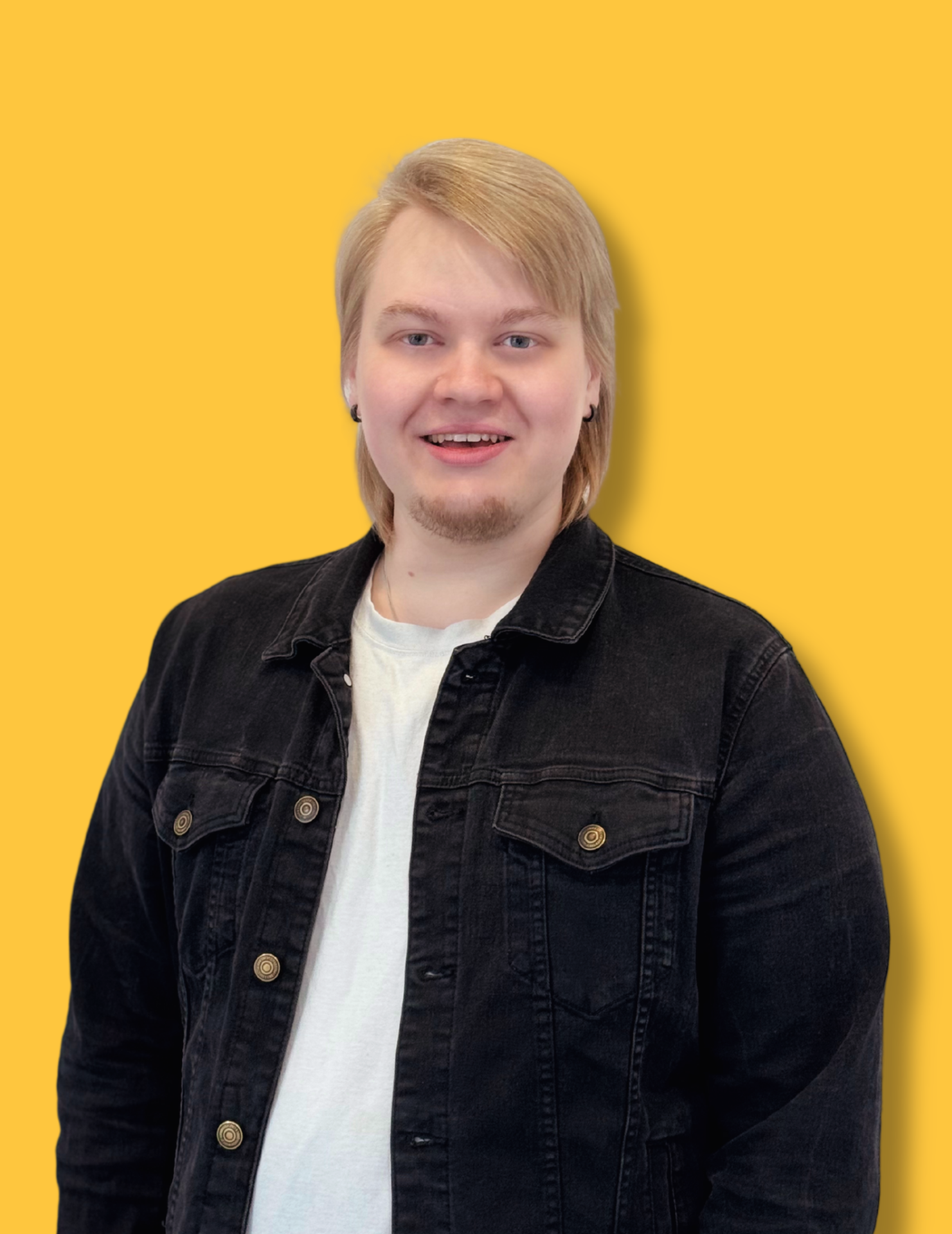 Vaaleahiuksinen mies hymyilee mustassa takissa ja valkoisessa paidassa kirkasta keltaista taustaa vasten.